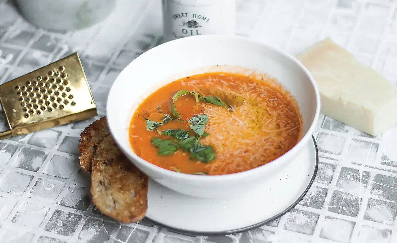 Aksamitna keto zupa pomidorowa z bazylią i parmezanem, podana z chrupiącym grzanką. Obok kawałek sera i złota tarka.