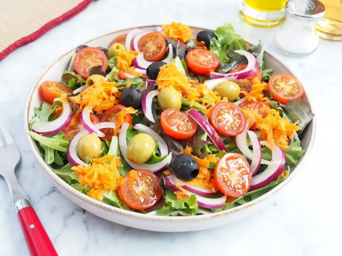 Ensalada mediterránea vibrante con tomates cherry, aceitunas, cebolla morada y zanahoria rallada sobre hojas verdes frescas.
