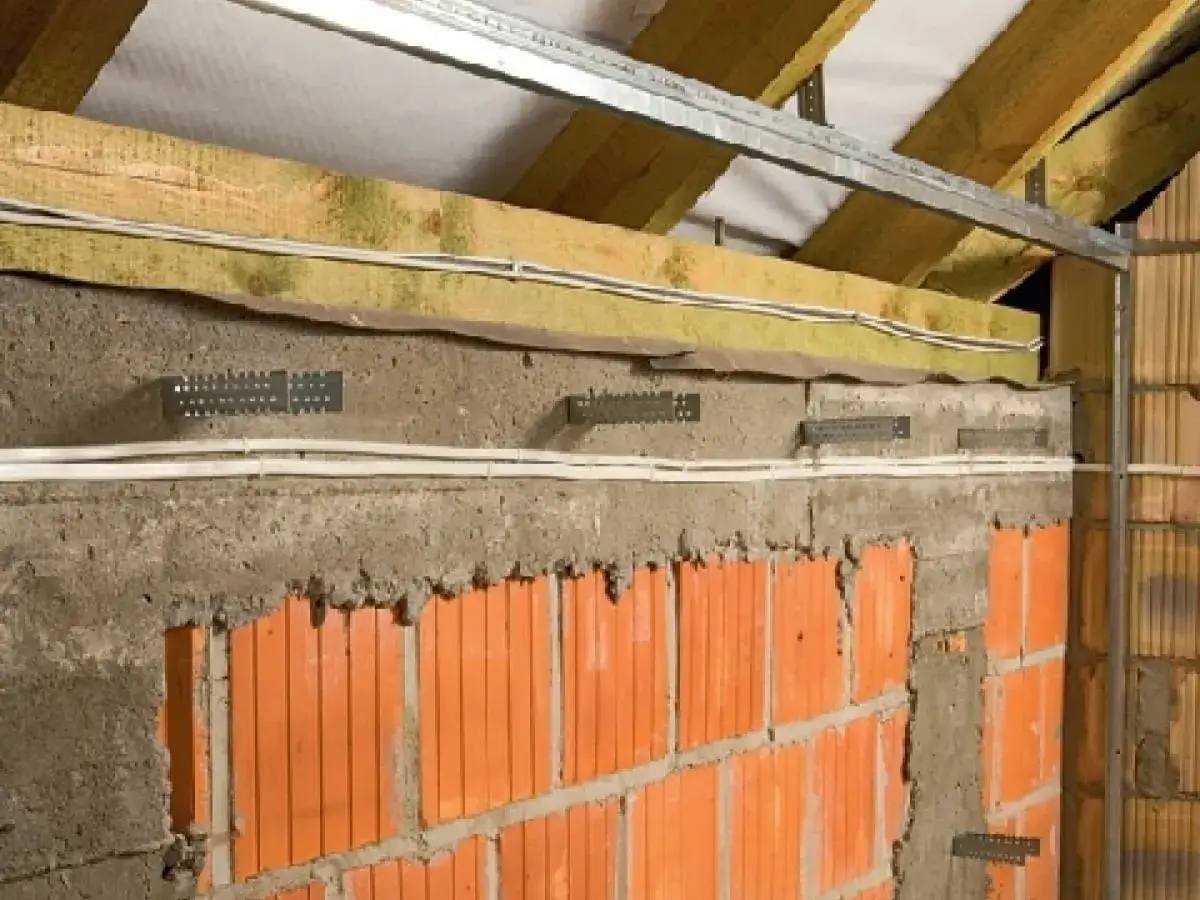 Budowa ścianki kolankowej w trakcie. Widoczna cegła, beton, izolacja i przewody elektryczne. Metalowe uchwyty do mocowania.