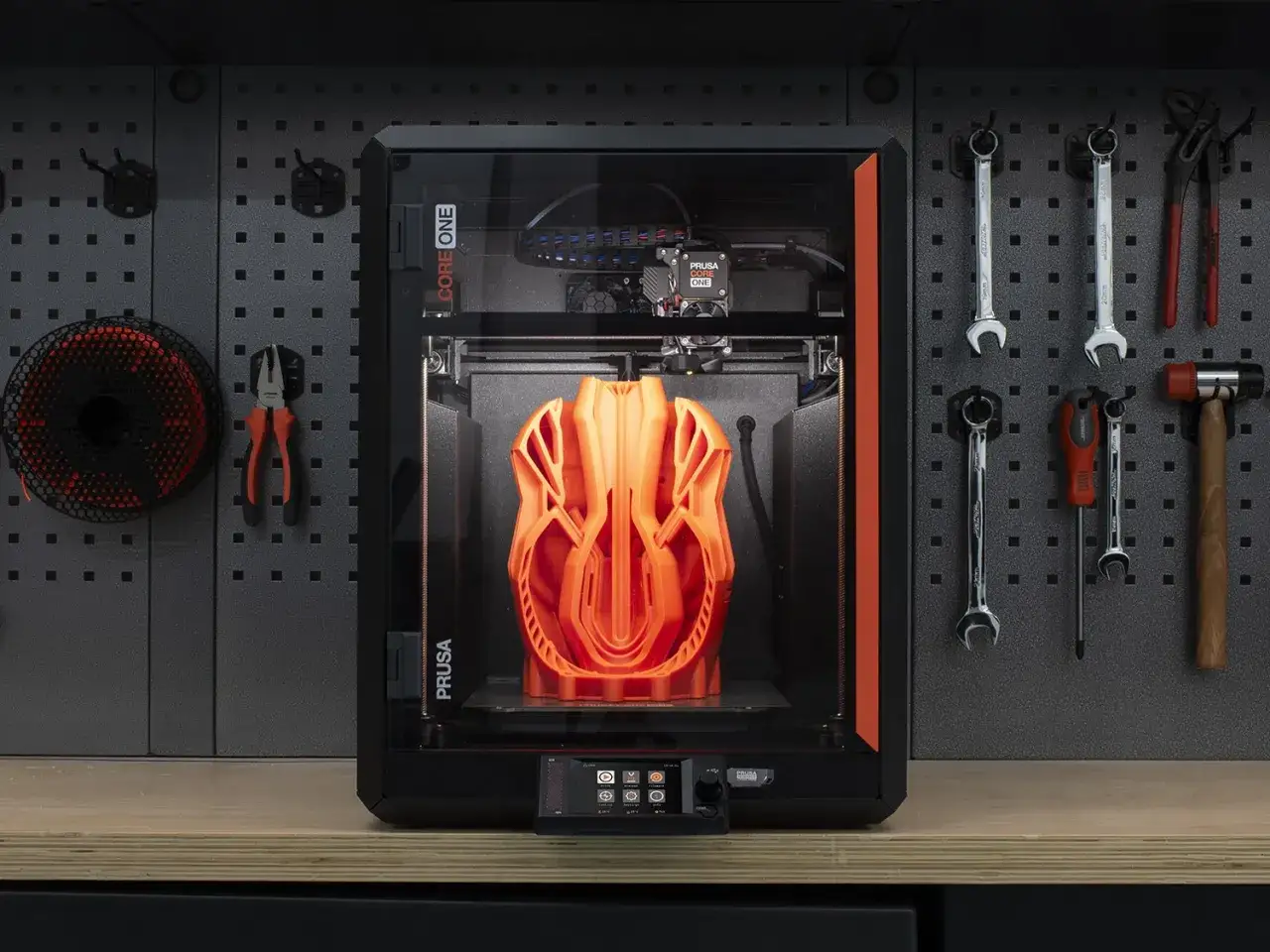 Pomarańczowy wydruk 3D w drukarce Prusa Core One. Obok wiszą narzędzia: klucze, młotek, kombinerki i szpulka filamentu.