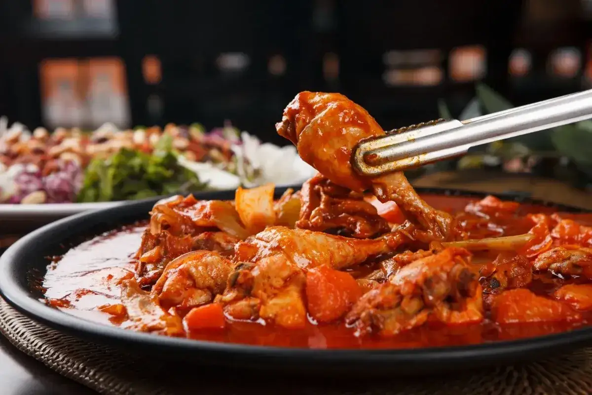 Un succulento friccò di pollo in una salsa rossa piccante, con pezzi di verdura. Una pinza solleva un pezzo di pollo.