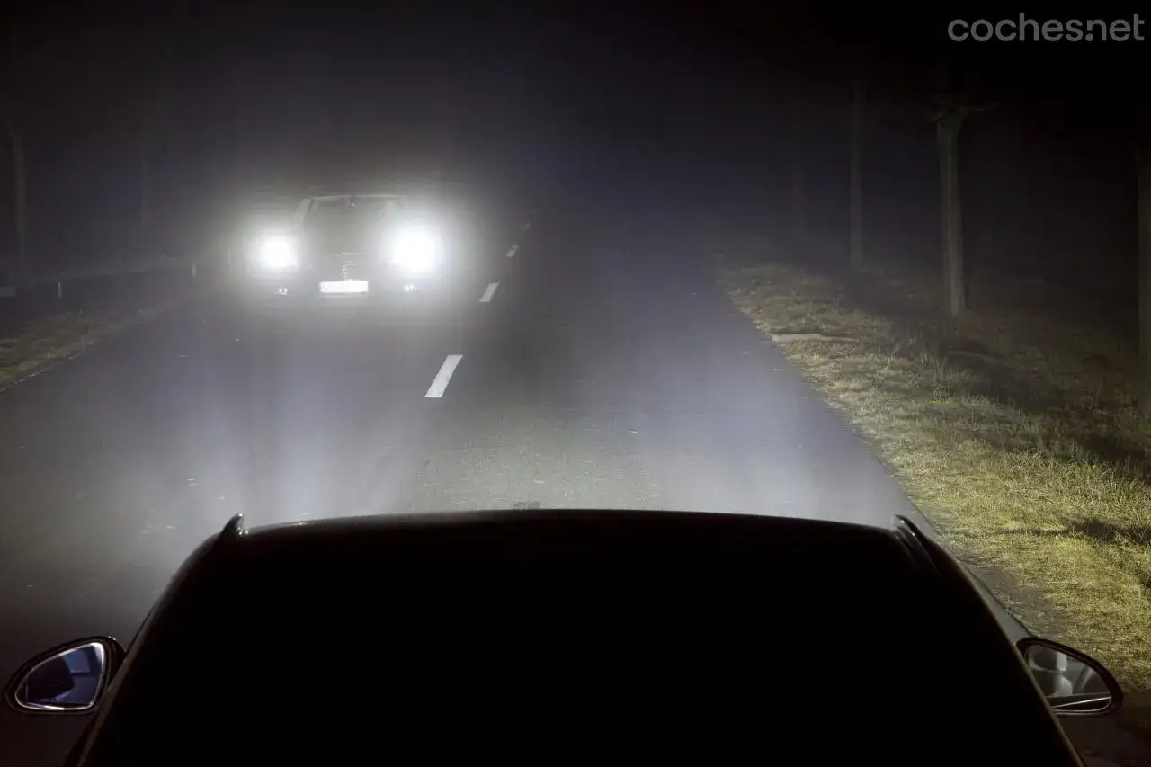 Luces largas iluminan una carretera oscura y neblinosa. Un coche se acerca, sus faros brillan intensamente.