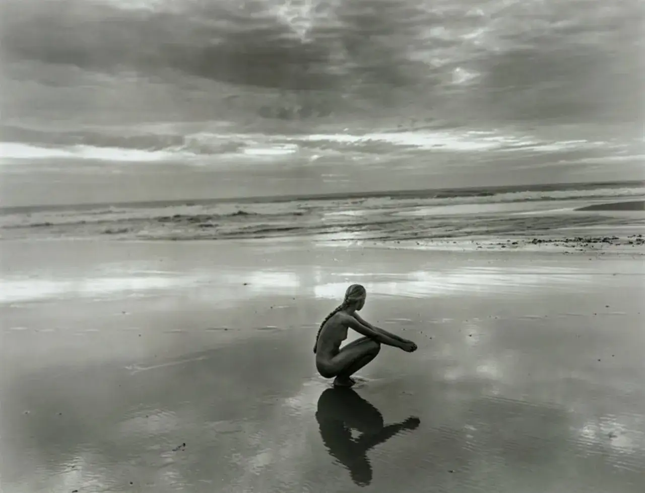 A young woman with a braid squats on a wet beach, her reflection mirroring her pose. The Jock Sturges style captures the serene fanny against a dramatic, cloudy sky and ocean backdrop.