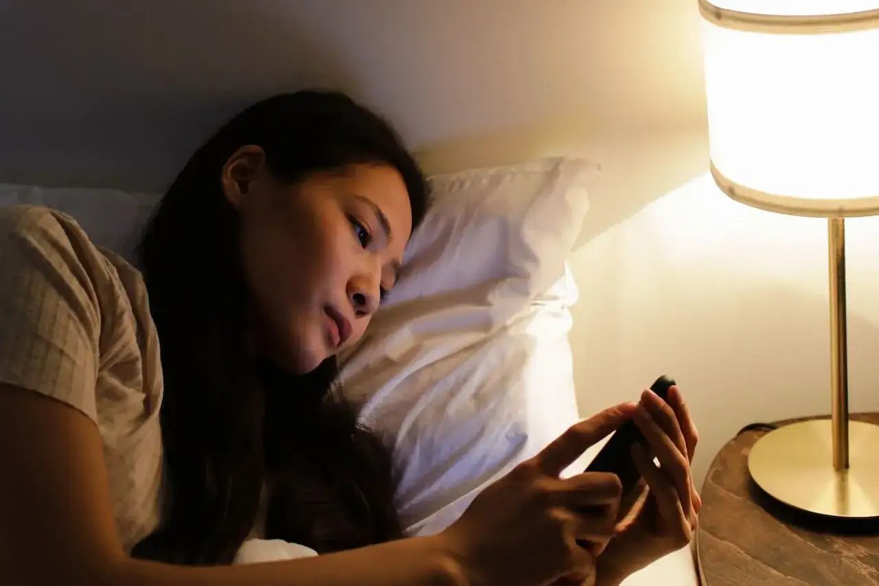 Junge Frau liegt im Bett, schaut auf ihr Handy. Das Licht der Nachttischlampe wirft einen warmen Schein. Vielleicht lernt sie für eine CBT-Prüfung.