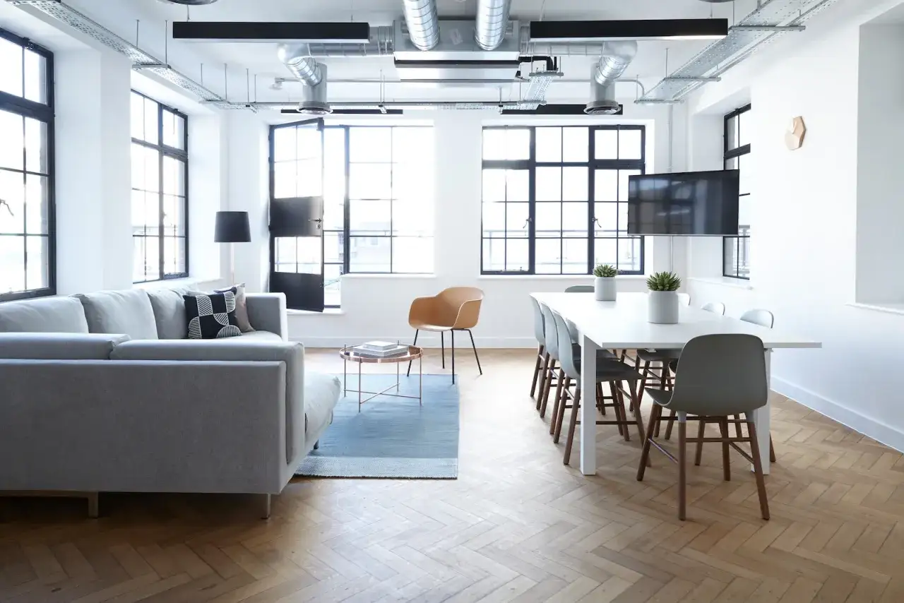 Nowoczesne mieszkanie w lokalu użytkowym: szara sofa, stół z krzesłami, duże okna i widoczne instalacje.