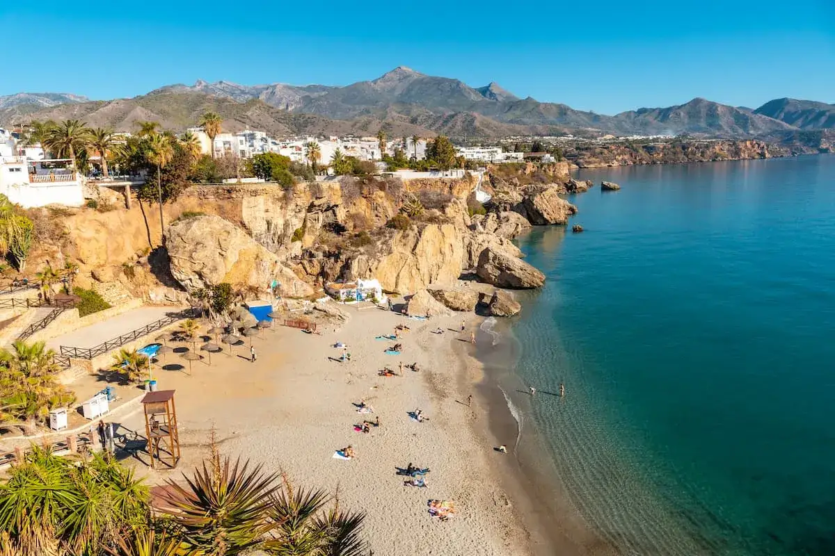 Playa con gente disfrutando del sol, acantilados y el mar azul, un paisaje típico de los pueblos costeros de Málaga.