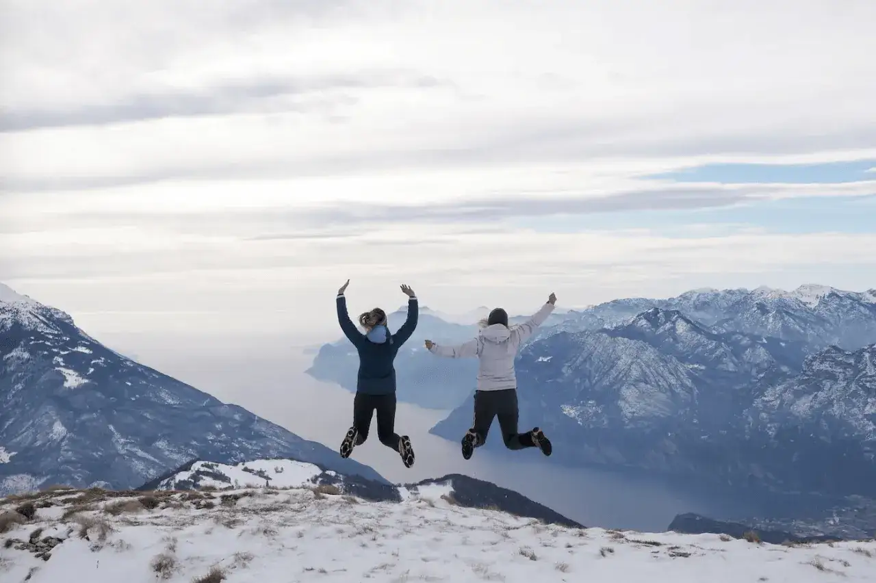 Due persone saltano con gioia su un crinale innevato, con il lago di Garda in inverno e le montagne sullo sfondo.