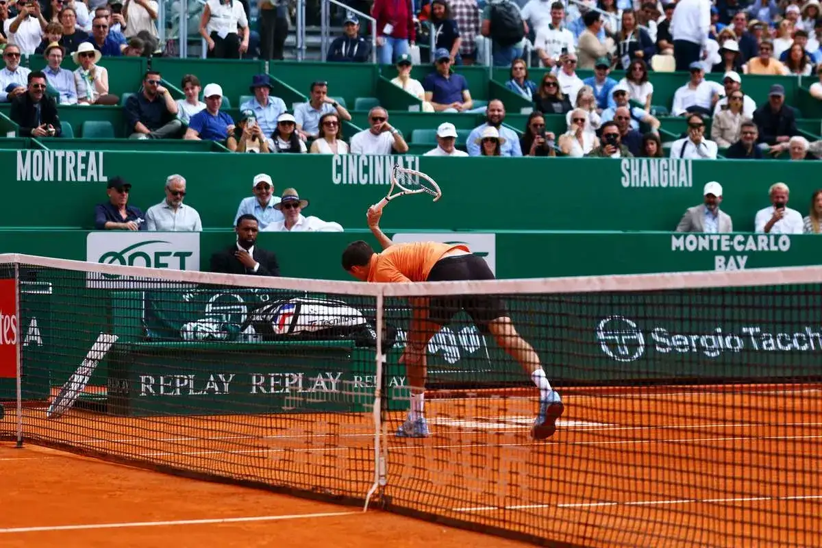 Gracz w pomarańczowej koszulce wykonuje serwis podczas meczu monte carlo tennis. Widownia obserwuje z trybun.