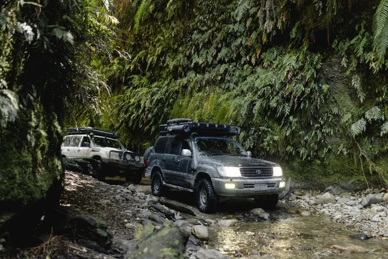 Dwa samochody Toyota Land Cruiser 100 pokonują kamienisty strumień w gęstym, zielonym lesie.