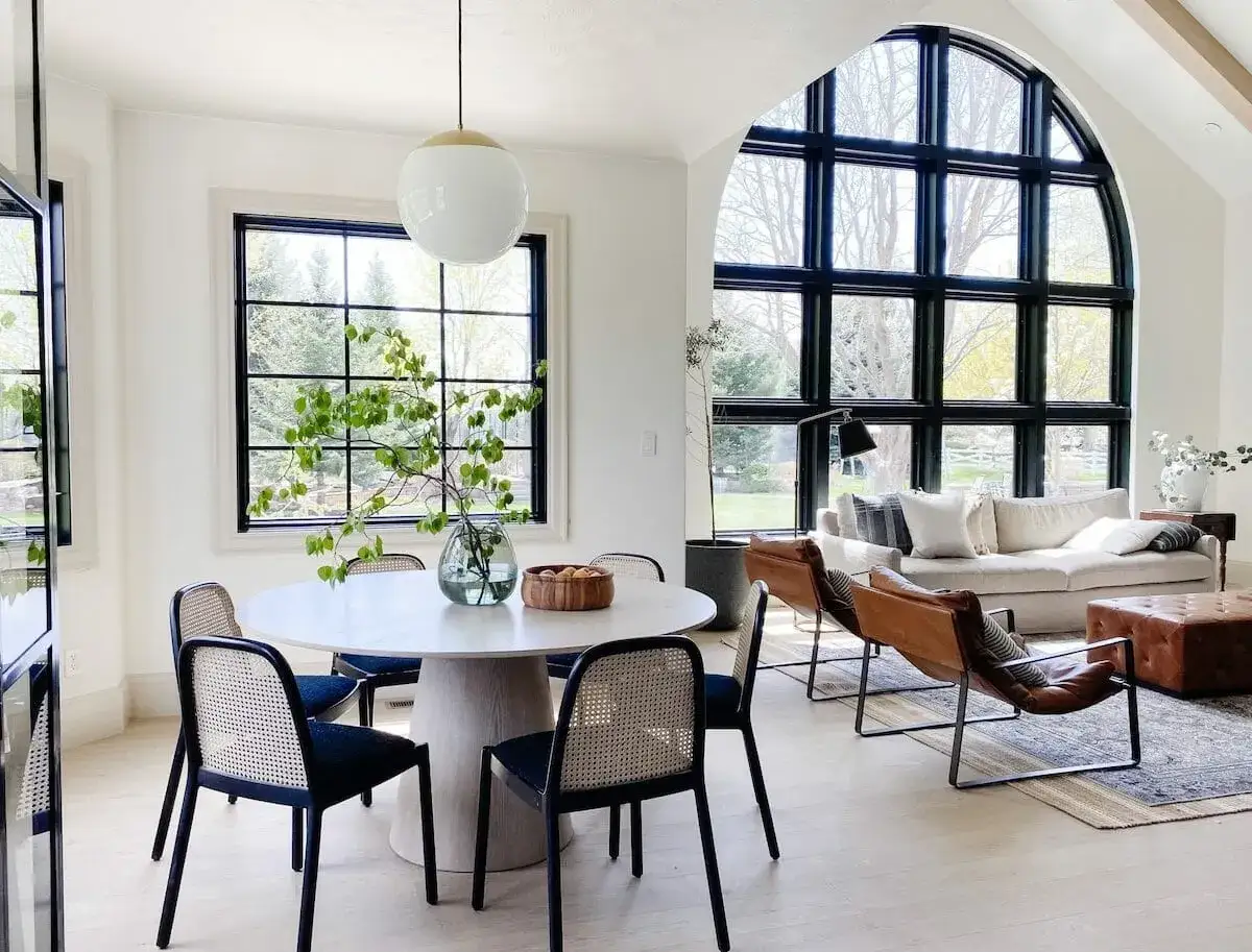 A bright, modern dining area with a round table and chairs, perfect for a breakfast nook built in. Large windows offer views of trees.