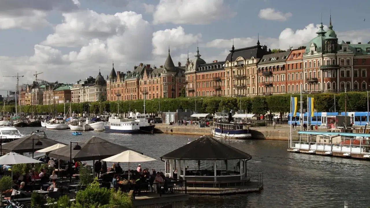 En vacker sommardag i Stockholm. Båtar ligger förtöjda vid kajen framför ståtliga byggnader. Folk sitter vid uteserveringar och njuter av utsikten.