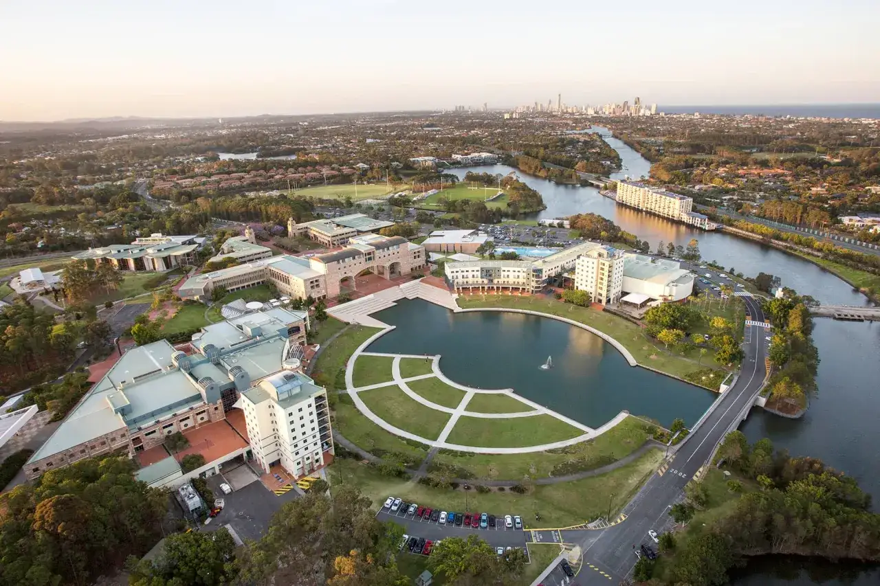 Luftaufnahme des Campus der Bond University mit einem zentralen See, umgeben von modernen Geb&auml;uden und Gr&uuml;nfl&auml;chen. Im Hintergrund erstreckt sich eine Stadtkulisse.