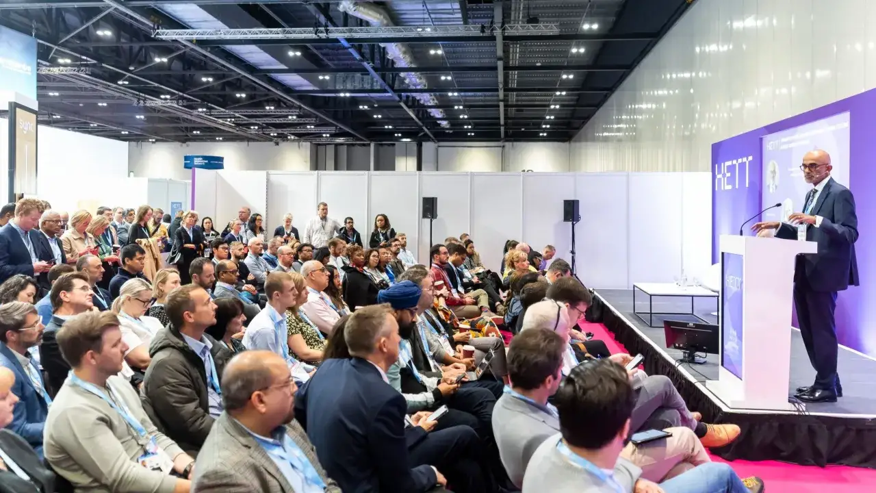 A speaker addresses a large audience at a conference. The audience, a good follower of the speaker's words, is seated and attentive.