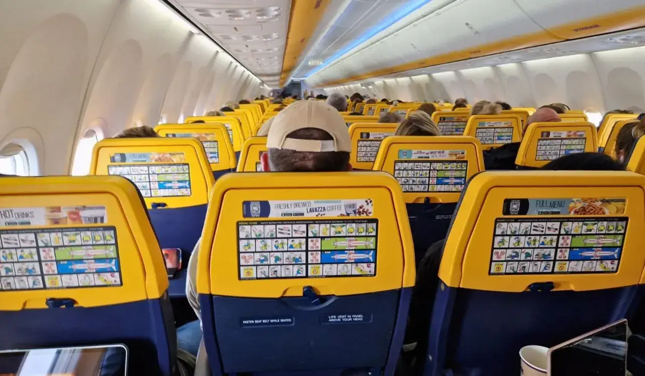 Widok na rząd żółtych oparć z ekranami w samolocie Ryanair. Widać pasażerów i instrukcje bezpieczeństwa. Idealne miejsca w samolocie Ryanair przy oknie.