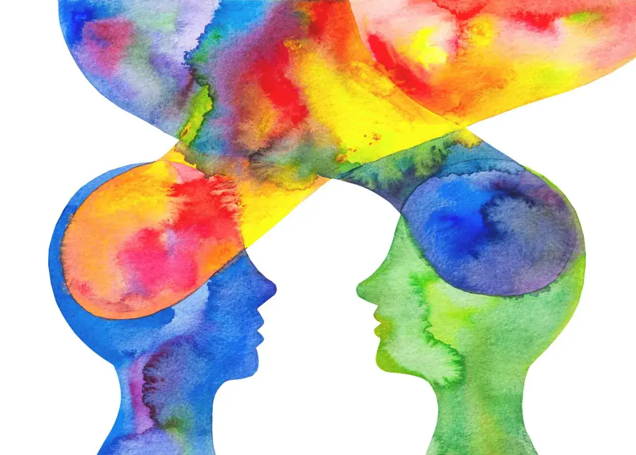 Zwei stilisierte Köpfe in Blau und Grün, die sich gegenüberstehen. Ihre Gedanken, dargestellt durch lebendige Farben, vermischen sich in der Mitte. Ein Symbol für die Vielfalt der **Charaktereigenschaften** und die Kraft der **Farben**.
