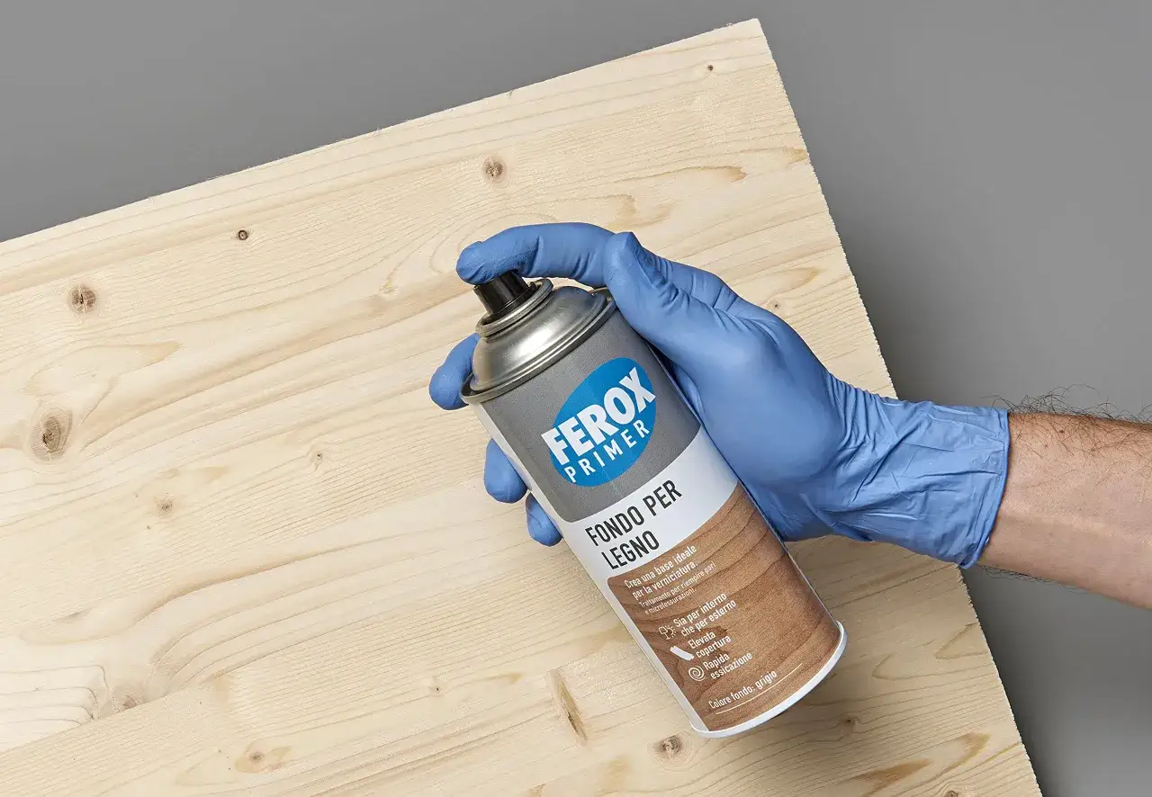 Mano con guanto blu tiene bomboletta di cementite per legno gi&agrave; verniciato, pronta per l'applicazione su una superficie grezza.