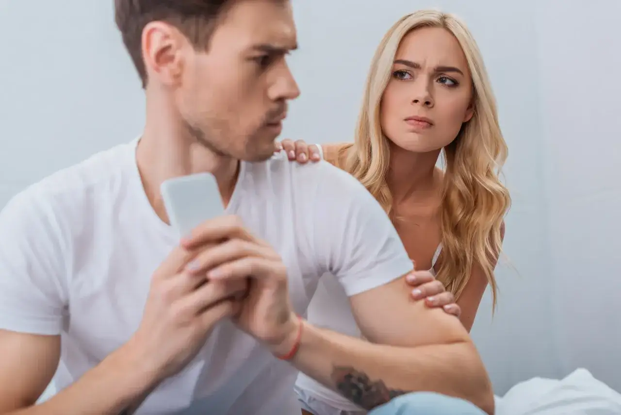Coppia con espressioni tese. Lei lo guarda con sospetto, lui è concentrato sul telefono, forse vittima di gelosia retroattiva.