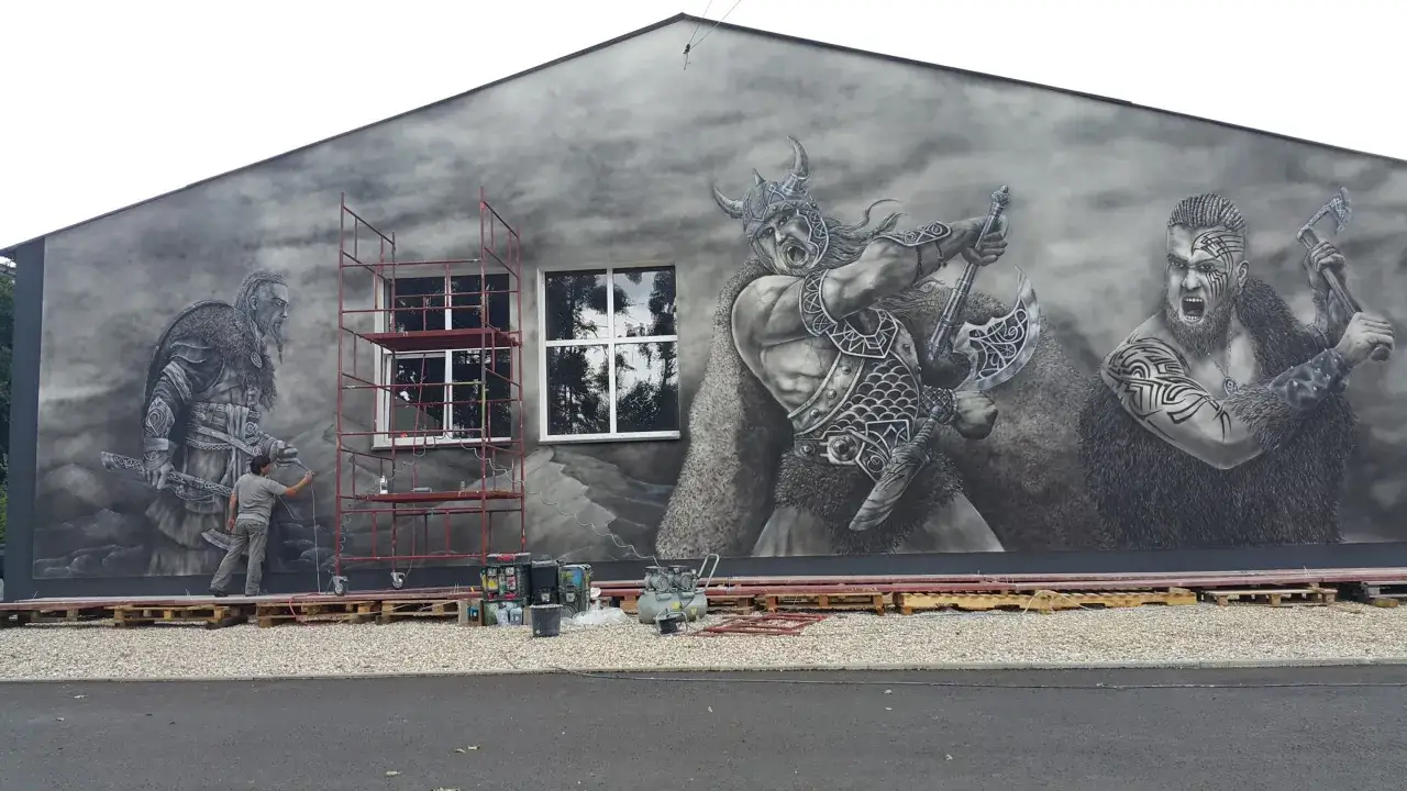 Mural na domu jednorodzinnym przedstawia trzech wojowników wikingów. Artysta w szarych barwach maluje ich na ścianie.