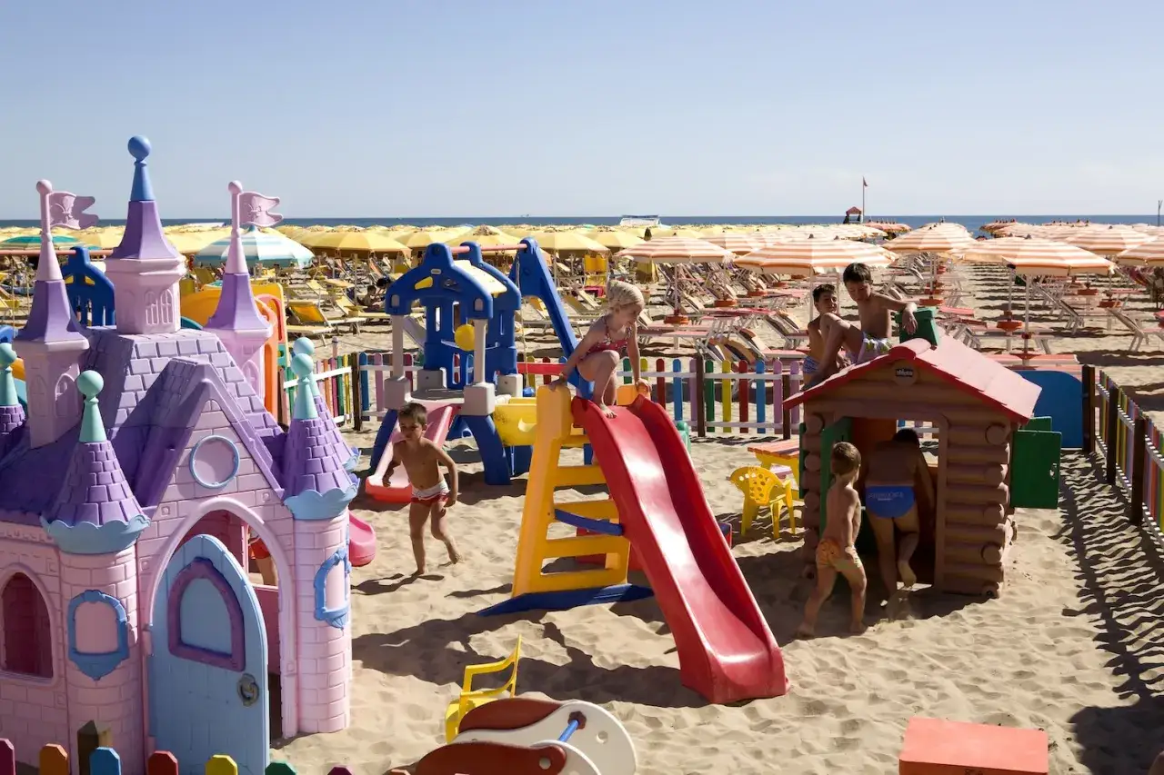 Bambini giocano felici sulla spiaggia della Riviera Romagnola, tra castelli colorati e scivoli, sotto il sole estivo.