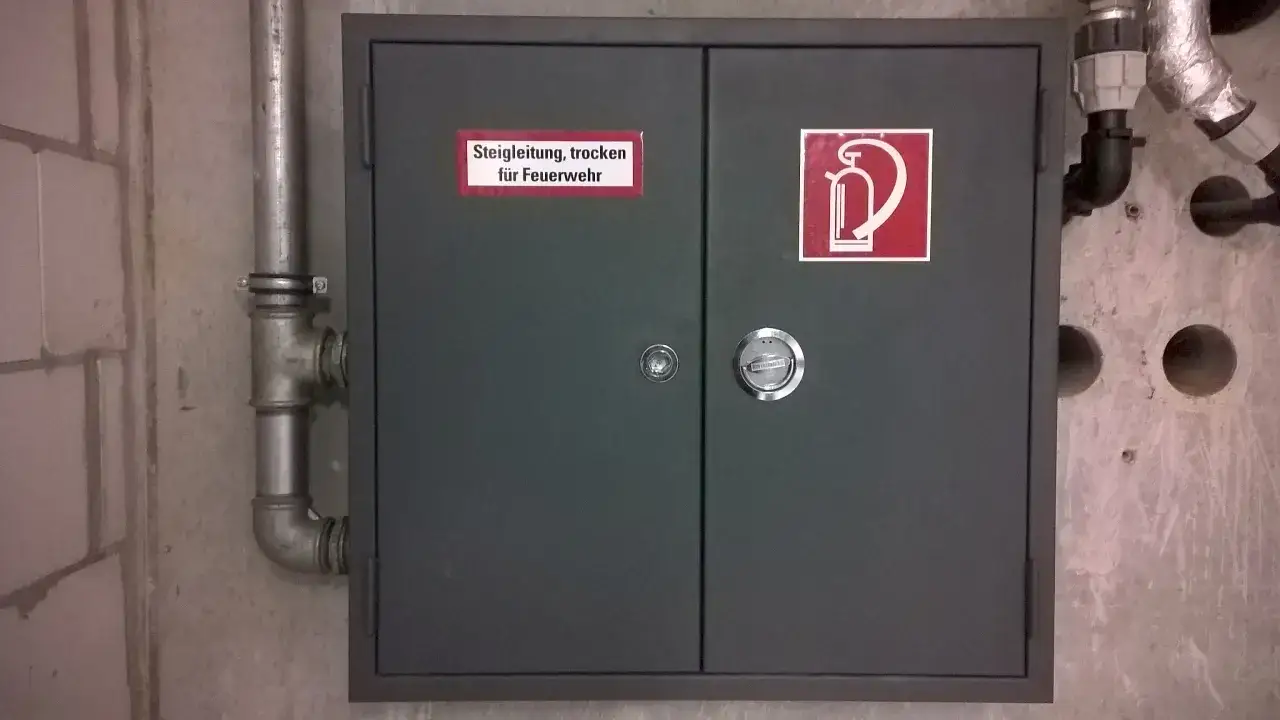 Grauer Schrank für Feuerwehr-Steigleitung, trocken, mit Brandschutzzeichen und DIN 14462-Kennzeichnung.