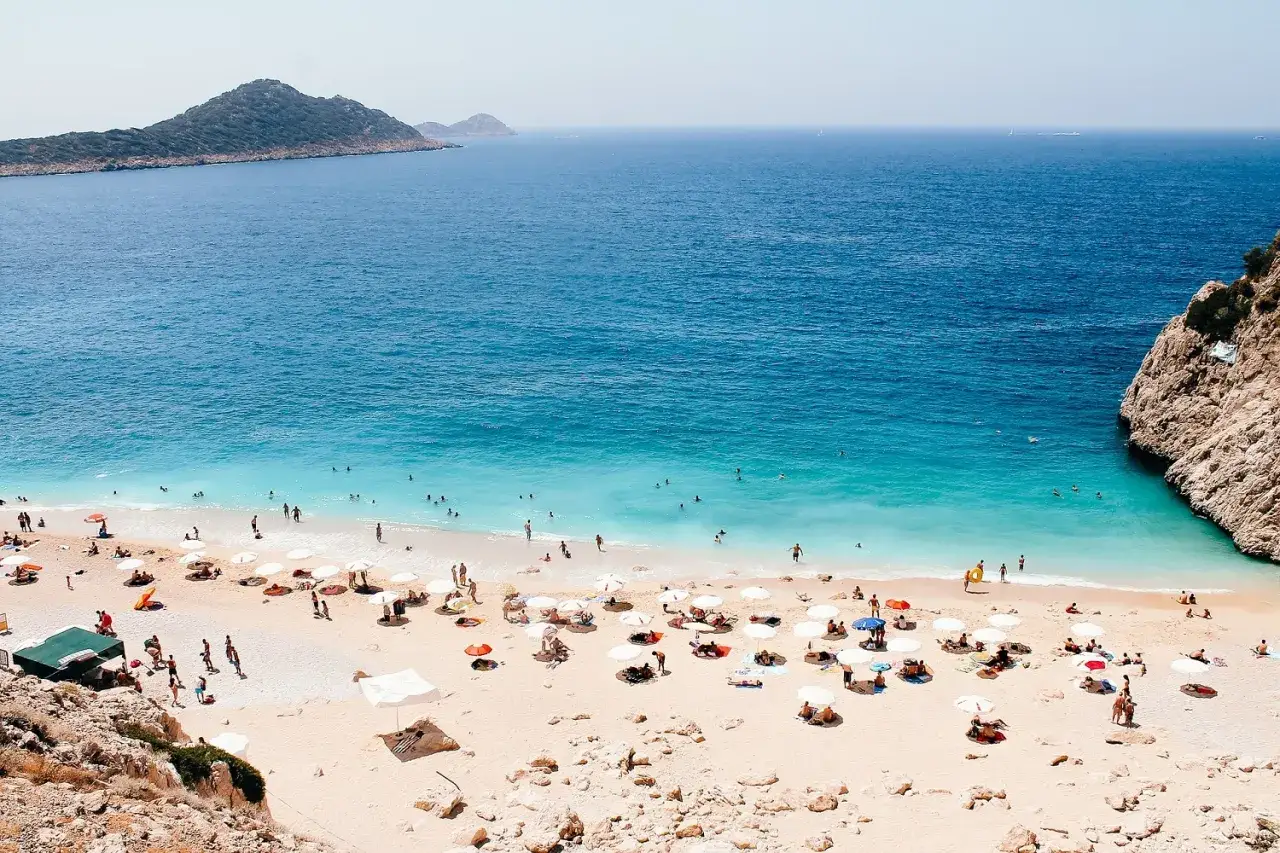 Piękna plaża z turkusową wodą, gdzie ludzie odpoczywają pod parasolami. Czy to Bodrum czy Alanya? Trudno powiedzieć, ale na pewno wakacyjny raj.