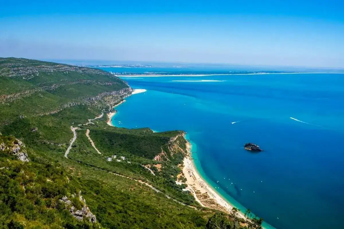 Atemberaubender Blick auf die K&uuml;ste von Set&uacute;bal, Portugal, mit &uuml;ppig gr&uuml;nen H&uuml;geln, die ins t&uuml;rkisfarbene Meer abfallen.