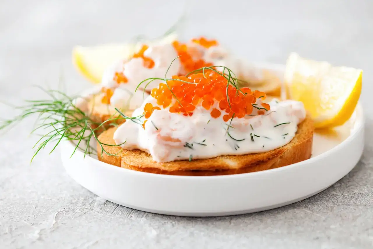 H&auml;r &auml;r godaste toast skagen p&aring; en vit tallrik, toppad med r&auml;kor, majonn&auml;s, dill och rom. Citronklyfta och dillkvistar som dekoration.