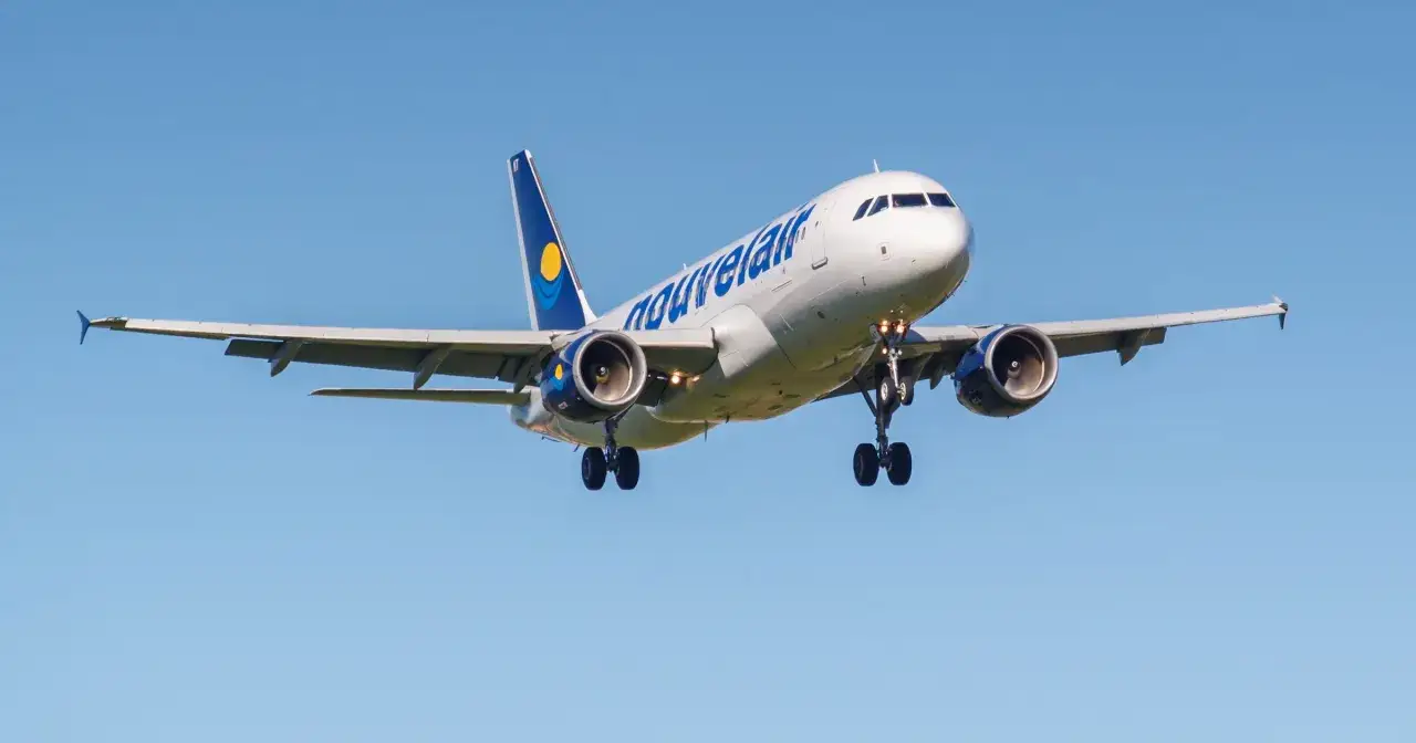 Samolot linii nouvelair z wysuniętym podwoziem zbliża się do lądowania na tle błękitnego nieba.