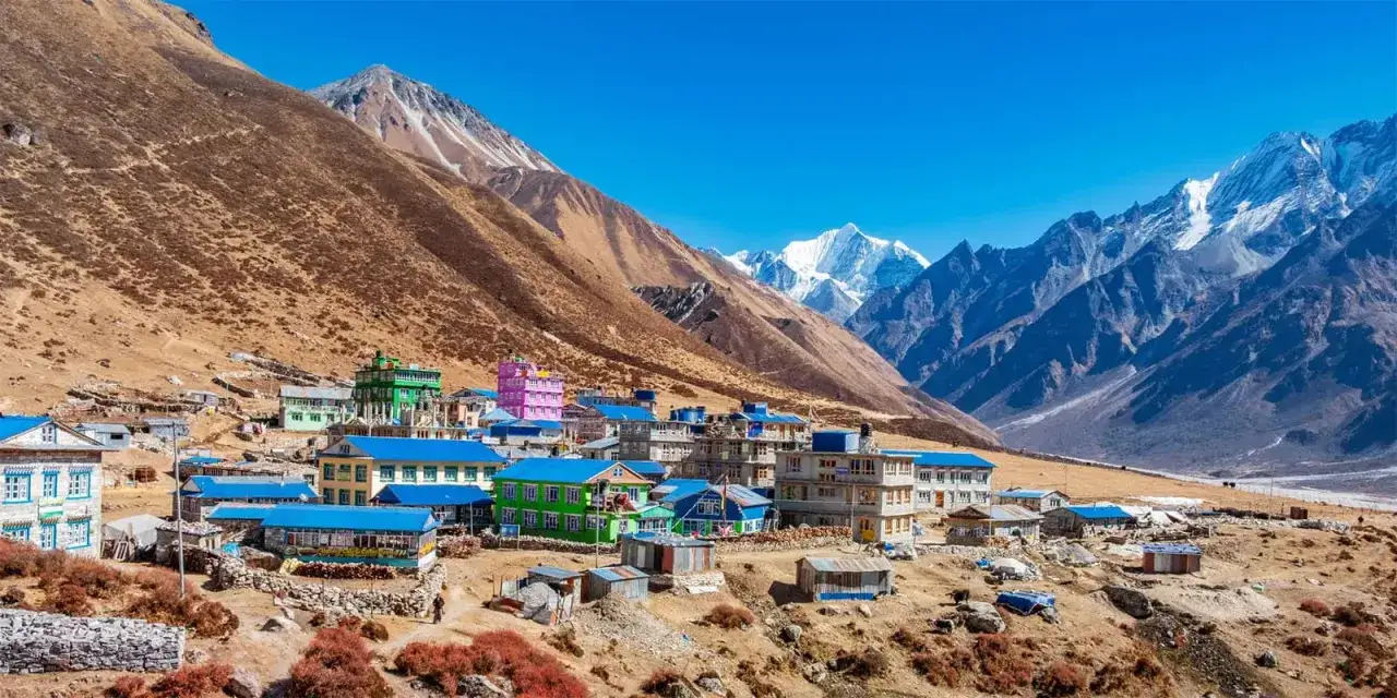 Villaggio colorato ai piedi dell'alta montagna himalayana, con case dai tetti blu e montagne innevate sullo sfondo.