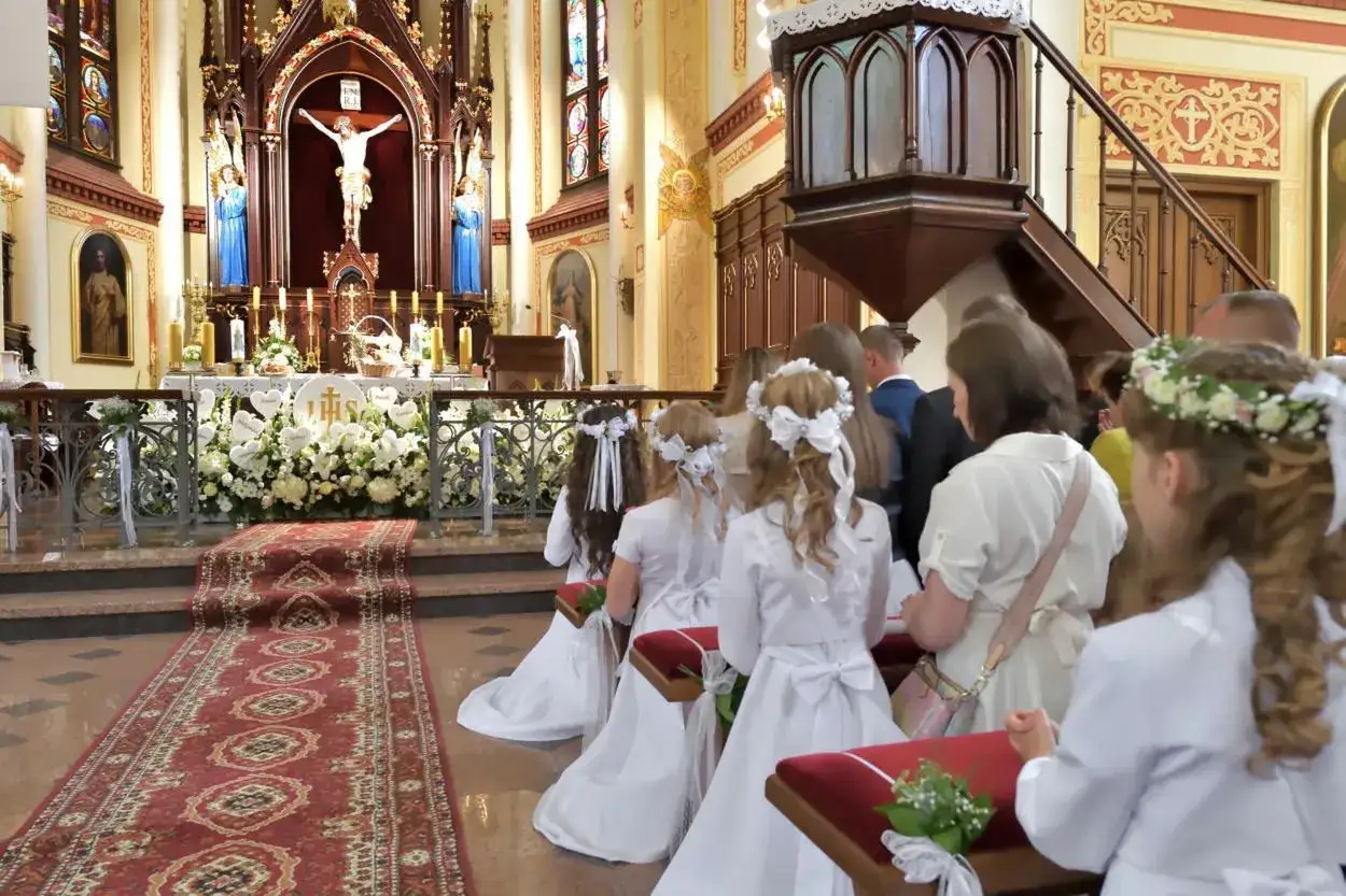 Dzieci w białych strojach komunijnych klęczą przed ołtarzem. Wnętrze kościoła, bogato zdobione, sugeruje uroczystość, która może różnić się w szczegółach liturgii między kościołem katolickim a prawosławnym.