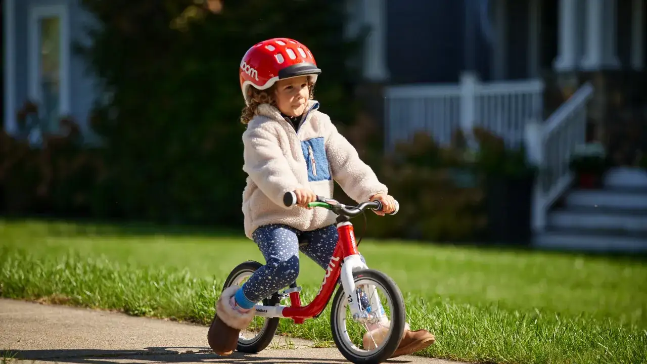 A child in a red Woom helmet rides a red woom balance bike on a sunny day.