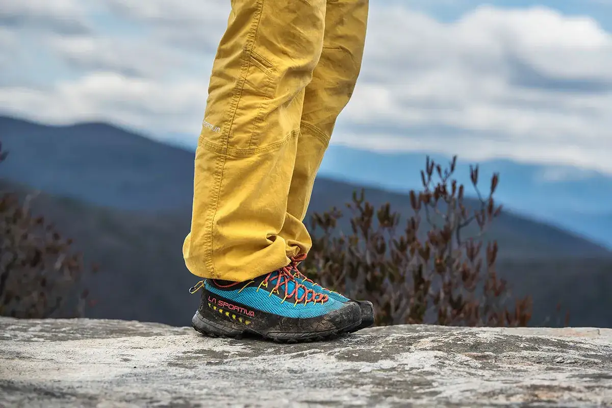 A person stands on a rock, wearing yellow pants and blue approach vs hiking shoes.