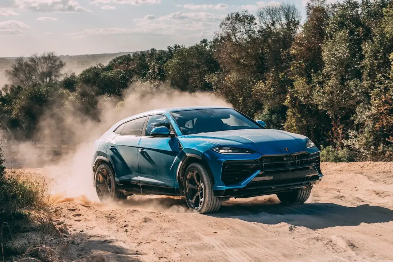 Niebieski Lamborghini Urus szaleje po piaszczystej drodze, wzbijając tumany kurzu.