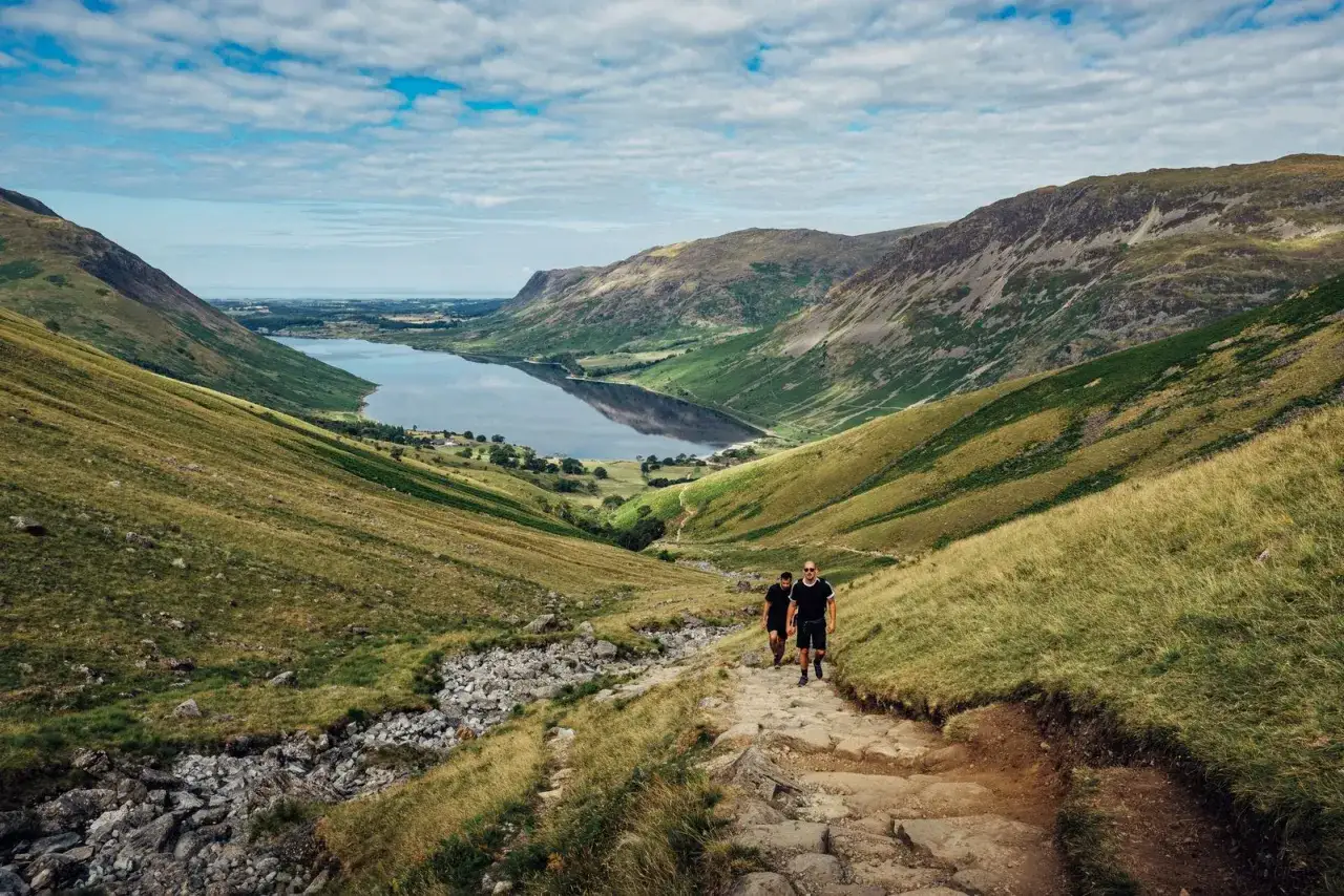Zwei Wanderer steigen einen steinigen Pfad hinab, mit Blick auf einen ruhigen See im Lake District.