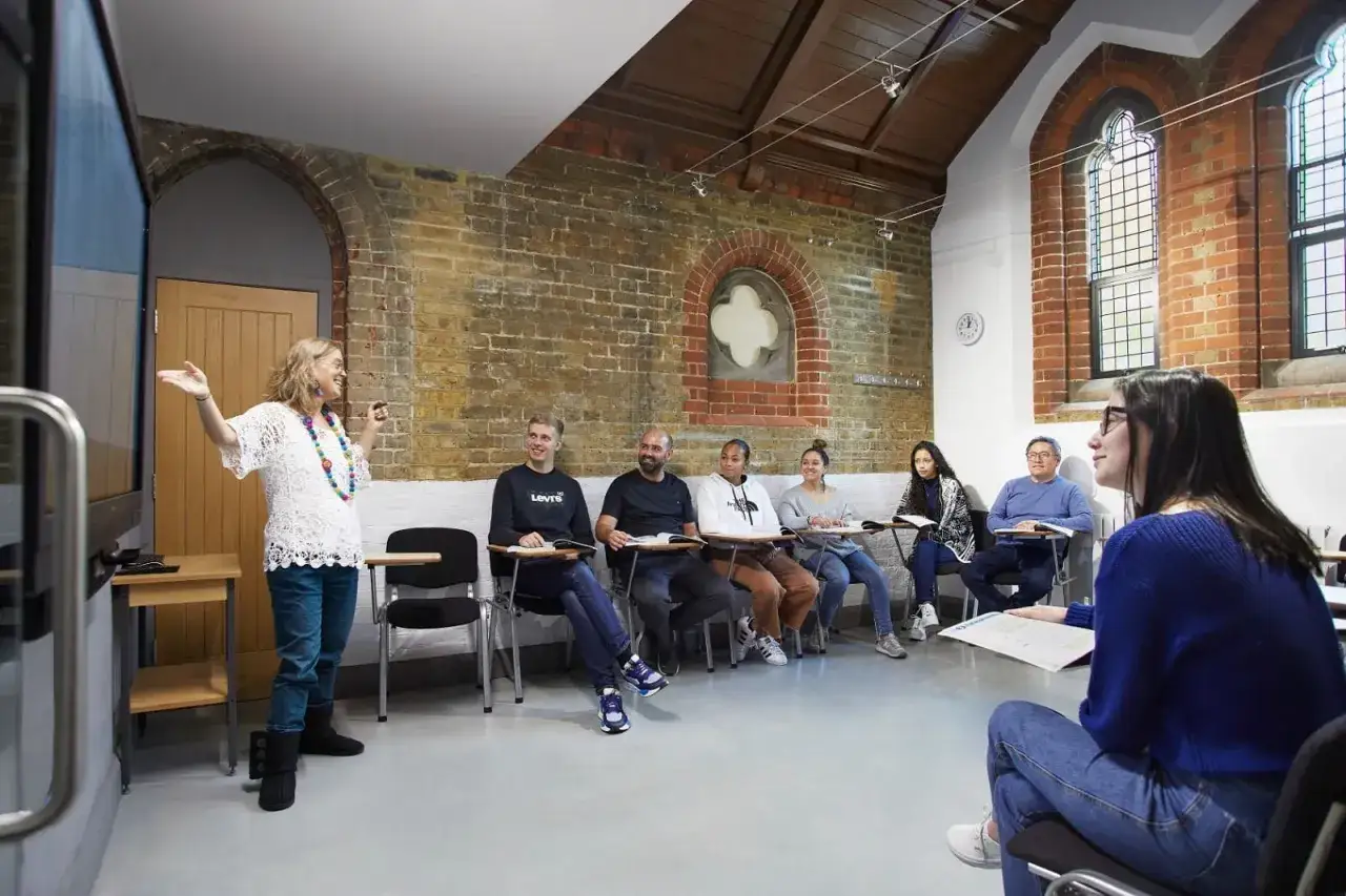 Clase de inglés en Londres. Un grupo diverso de estudiantes escucha atentamente a su profesora, quien gesticula animadamente frente a una pantalla.
