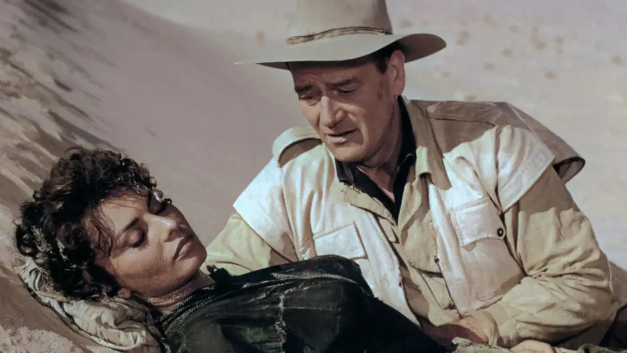 John Wayne troszczy się o swoją żonę, kt&oacute;ra leży wyczerpana na piasku.