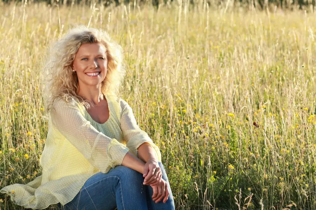 Uśmiechnięta kobieta po 50. siedzi na łące pełnej wysokich traw i ż&oacute;łtych kwiat&oacute;w. Jestem samotna po 50. i cieszę się chwilą spokoju.