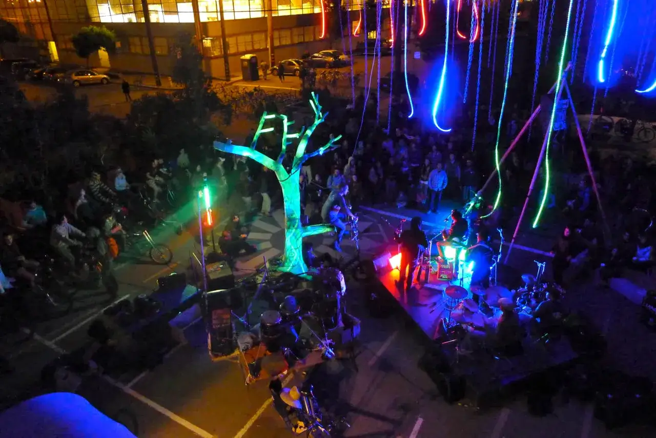 Un colorato teatro a pedali con un albero luminoso e musicisti, illuminato da luci al neon blu e verdi, con una folla che osserva.