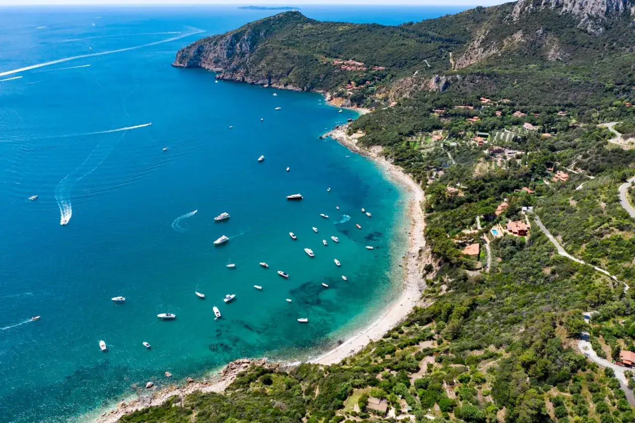 Costa dell'Argentario: cosa vedere? Baia con acque cristalline, spiagge dorate e ville immerse nel verde. Barche a vela e yacht solcano il mare blu.