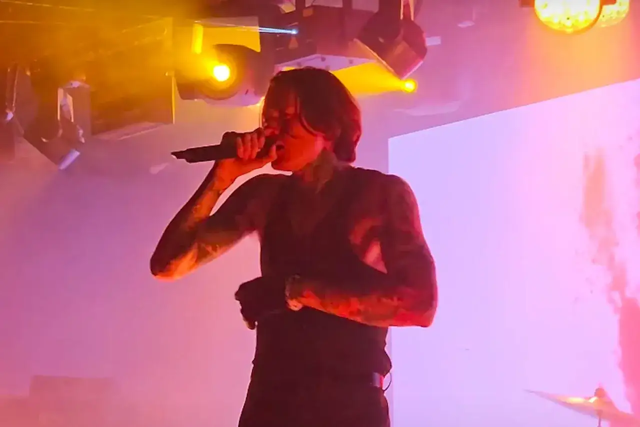 Noah Sebastian, tattooed and intense, belts into a microphone on a smoky stage, bathed in vibrant pink and yellow lights.