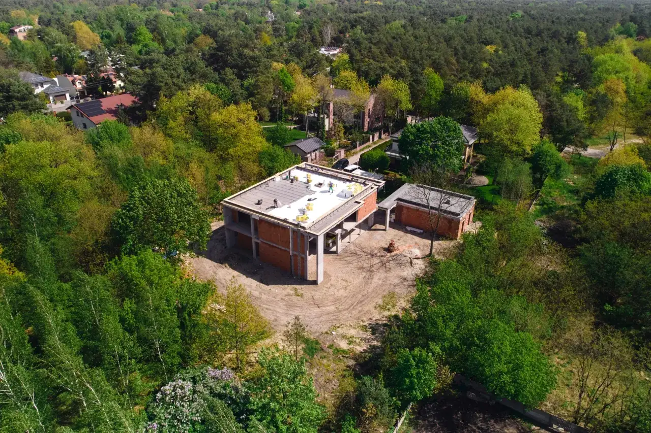 Budowa domu w Warszawie: stan surowy otwarty, prace na dachu. Wokół zieleń i inne domy.