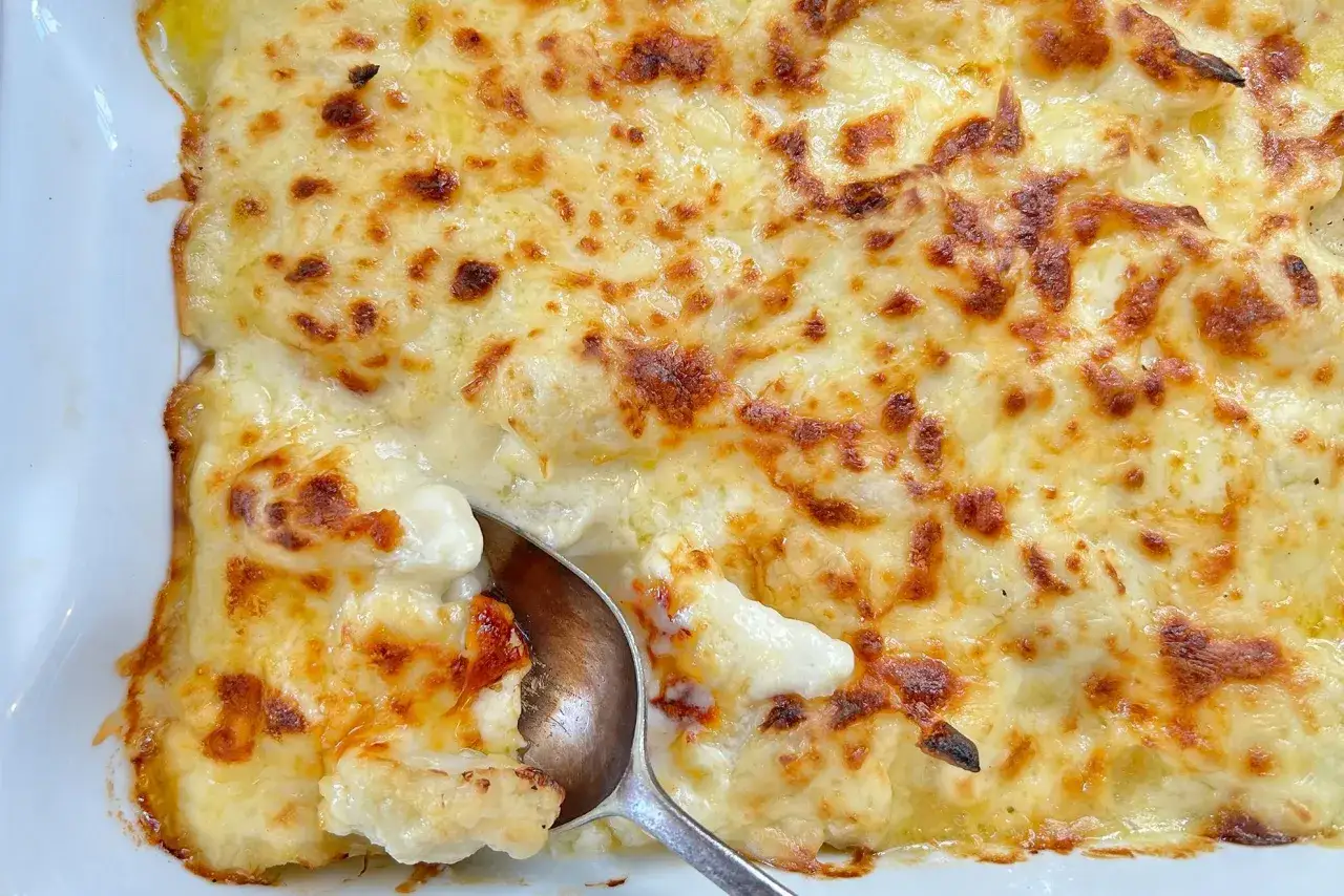 Coliflor gratinada con bechamel cremosa y queso dorado. Una cuchara sirve una porción de este delicioso plato.