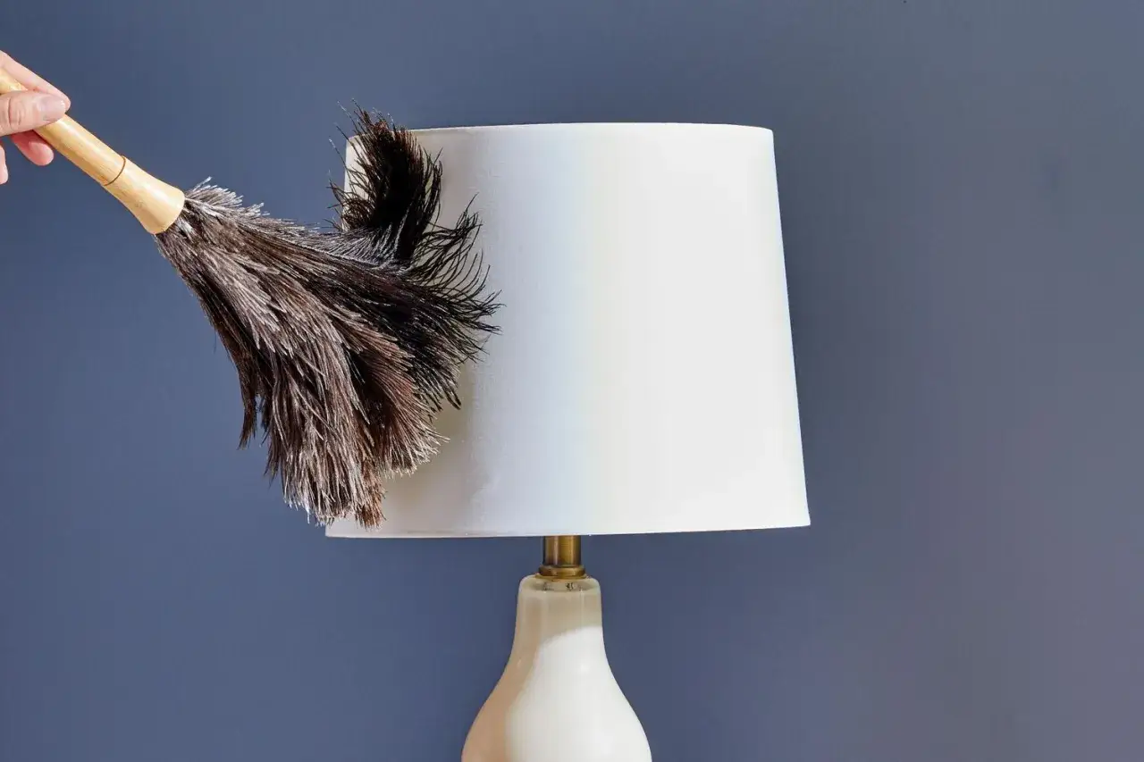 A hand uses a feather duster to gently clean a white lampshade, showing how to clean lamp shades effectively.