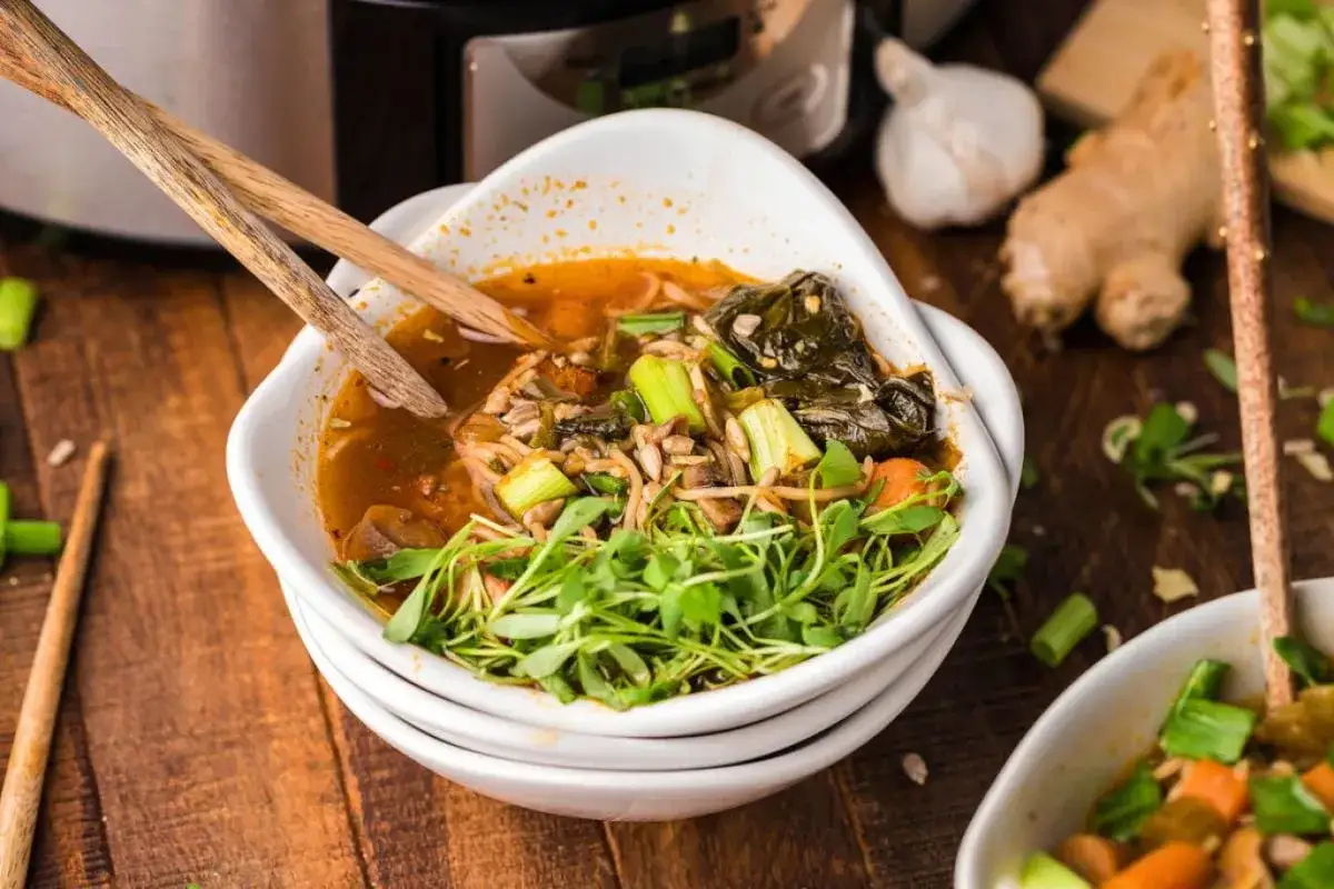 Un bol de ramen veggie fumant, garni de pousses fraîches, de céleri et de légumes verts. Des baguettes en bois reposent sur le bord.