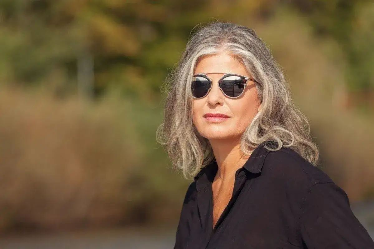 Donna con eleganti capelli bianchi e occhiali da sole, con uno sfondo sfocato di alberi autunnali.