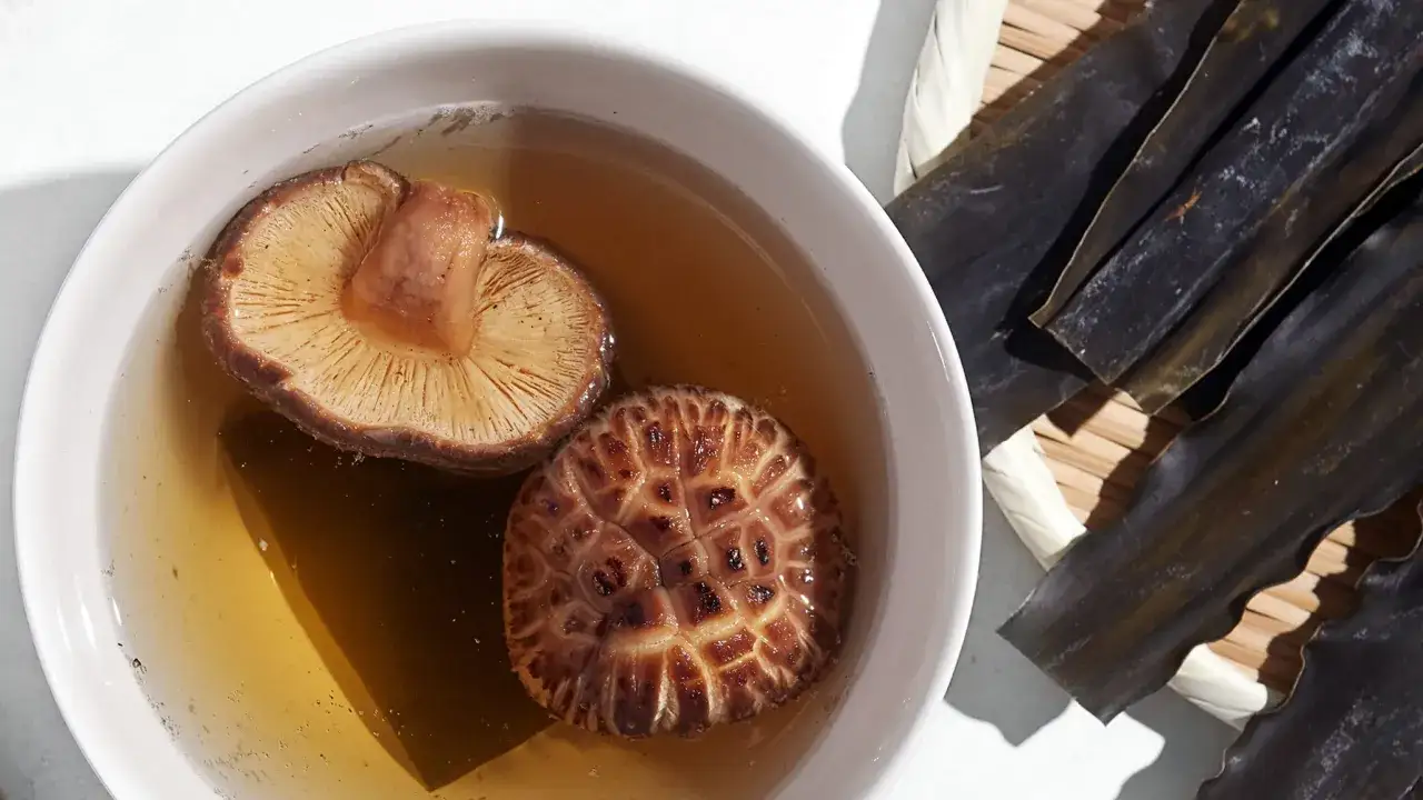 Two shiitake mushrooms steep in a bowl of golden liquid, creating a rich mushroom dashi. Dried kombu seaweed lies nearby.