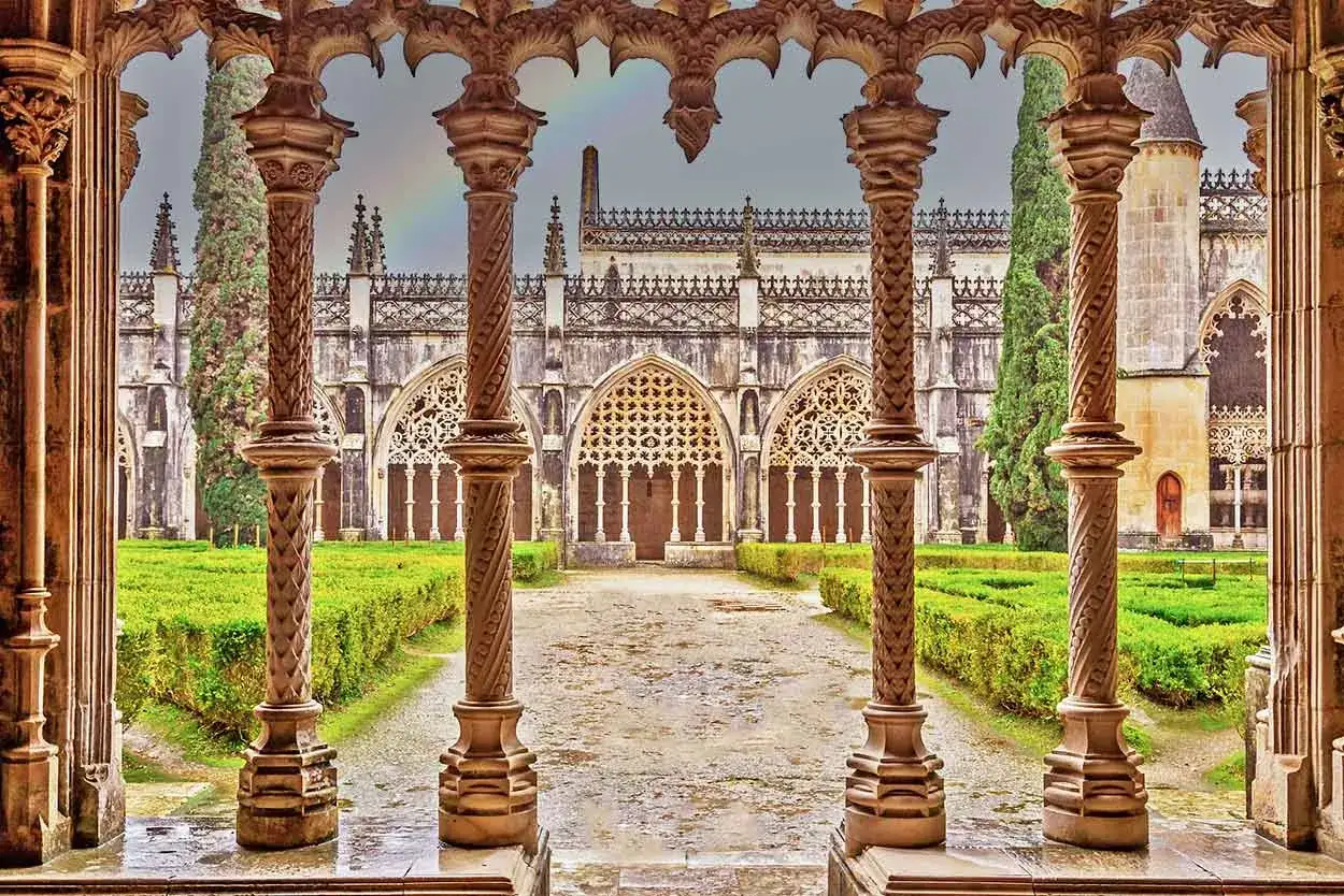 Ornate columns frame a view of the Batalha Monastery in Portugal, with manicured gardens and a cloudy sky.