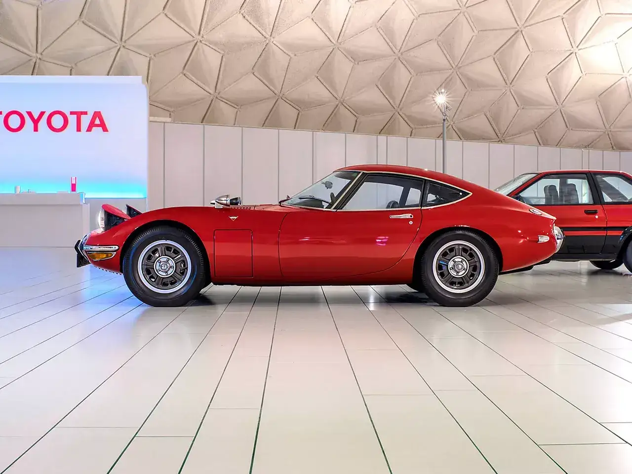 Klasyczny, czerwony Toyota 2000 GT z podniesionymi światłami, obok nowszego modelu.