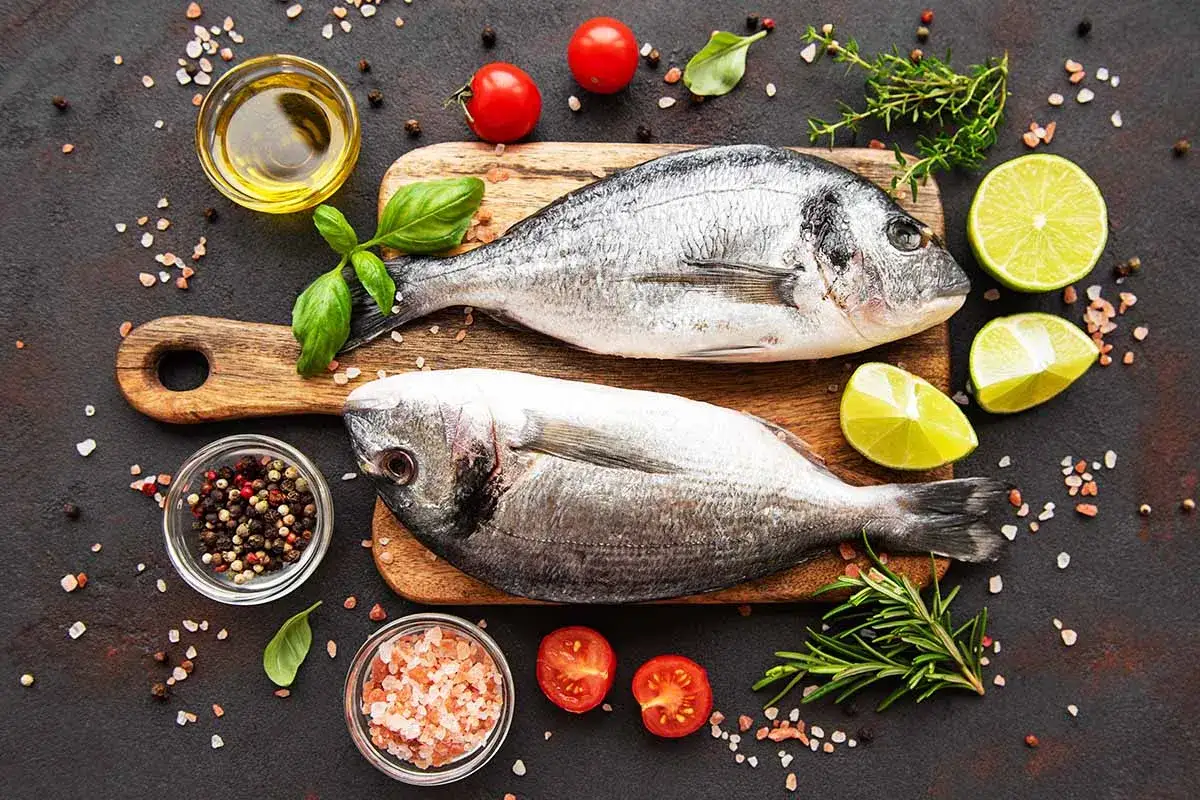 Deux poissons entiers prêts pour la cuisson, accompagnés de citrons, tomates, herbes et épices.