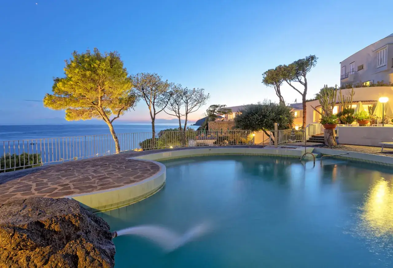 Piscina panoramica con vista mare al tramonto, alberi e l'edificio del hotel terme gran paradiso illuminato.